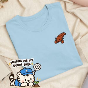 May include: A light blue t-shirt featuring a small embroidered brown lobster on the upper left chest. The shirt includes a graphic sticker of a white tiger wearing a blue cap, looking at a small sprout, with the text WAITING FOR MY DONUT TREE.