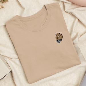 Könnte beinhalten: Ein beiges T-Shirt mit Rundhalsausschnitt und einer kleinen, gestickten Grafik eines braunen Quokkas auf der linken Brust. Das Quokka ist in sitzender Position mit zusammengelegten Pfoten und sichtbarem Schwanz in Braun-, Schwarz- und Grautönen dargestellt.