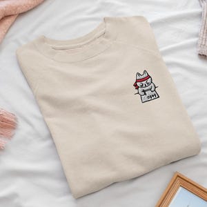 May include: A beige crewneck sweatshirt featuring a small embroidered graphic on the left chest. The embroidery depicts a white cat wearing a red headband, writing on a piece of paper with the text 1+1= on it. The sweatshirt has long sleeves.