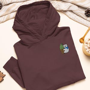 Op de afbeelding: Een donkerbruine hoodie met een geborduurde patch op de borst. De patch toont een witte ijsbeer met een blauwe sjaal die een groen softijsje in een bruin wafelhoorntje vasthoudt.