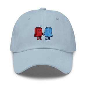 May include: A light blue baseball cap featuring an embroidered design of two small, anthropomorphic building blocks, one red and one blue, holding hands. The blocks have simple facial features, arms, and legs, centered on the front panel.