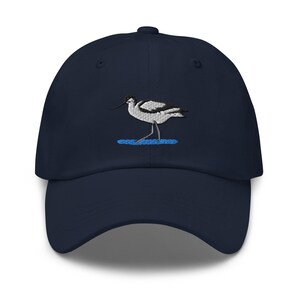 Op de afbeelding: Een marineblauwe baseballpet met een geborduurd ontwerp van een zwart-witte kluut die in een klein plasje blauw water staat. De pet heeft een gebogen klep, een gestructureerde kroon en een knoopje bovenop de stoffen panelen.