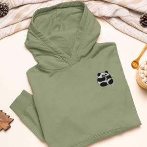 May include: A sage green hooded sweatshirt featuring a small embroidered graphic of a round black and white panda bear holding a green bamboo stalk on the left chest area. The hoodie includes a front kangaroo pocket and ribbed sleeve cuffs.