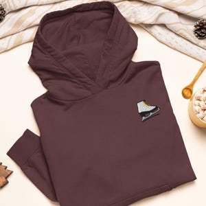 Könnte beinhalten: Ein dunkelbrauner Kapuzenpullover mit einer kleinen Stickerei auf der Brust. Die Stickerei zeigt einen weißen Schlittschuh mit schwarzer Kufe und gelben Schnürsenkeln. Der Hoodie hat lange Ärmel mit gerippten Bündchen und eine große Vordertasche.