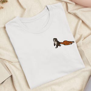 Op de afbeelding: Een wit t-shirt met korte mouwen en een kleine geborduurde afbeelding op de linkerborst. Het ontwerp toont een kleine primaat met donkere vacht die een grote, getextureerde oranje wortel met zijn handen over de stof trekt.