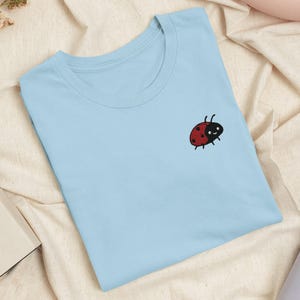 Può includere: Una t-shirt azzurro chiaro con una piccola coccinella ricamata sulla parte superiore sinistra del petto. La coccinella è rossa con puntini neri, testa nera e due antenne, raffigurata con un volto semplice e sorridente.