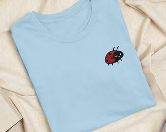 Puede incluir: Una camiseta de cuello redondo en color azul claro con una pequeña mariquita bordada en el pecho izquierdo. La mariquita tiene alas rojas con puntos negros, cabeza negra y una cara sencilla y sonriente con dos antenas.