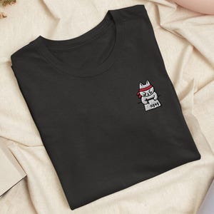 May include: A black crew neck t-shirt featuring a small embroidered graphic on the chest. The design depicts a white cat wearing a red headband, focused on writing on a piece of paper that displays the mathematical equation 1+1=2 in black ink.