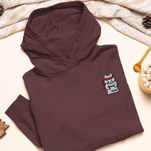 May include: A dark maroon hooded sweatshirt featuring an embroidered graphic on the left chest. The design depicts a white cat wearing a red headband, leaning over a gray desk while writing the equation 1+1 on a piece of paper with a pencil.