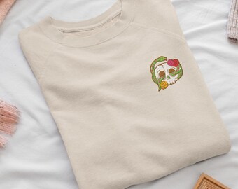 May include: A beige crewneck sweatshirt featuring a small embroidered design on the upper left chest. The embroidery depicts a white skull intertwined with a green snake, accented by a red rose and a yellow rose near the skull.