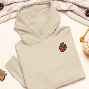 May include: A beige hooded sweatshirt featuring a small, embroidered strawberry patch on the left chest. The strawberry is red with a green stem and yellow seeds, stitched with a black outline. The hoodie has long sleeves with ribbed cuffs.