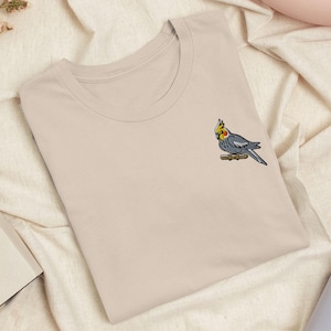 Puede incluir: Una camiseta beige de cuello redondo con un pequeño bordado en la parte superior izquierda del pecho. El diseño muestra una cacatúa ninfa gris con cresta amarilla y manchas naranjas en las mejillas, posada sobre una rama marrón.