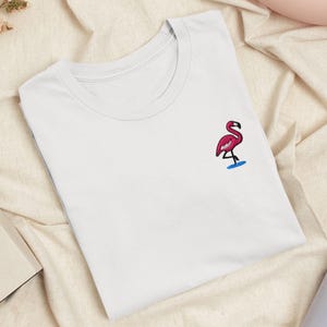 以下が含まれることがあります： 胸元に小さな刺繍が施された白い半袖Tシャツ。刺繍は、片足で立つ鮮やかなピンク色のフラミンゴが、水を表現した小さな青い楕円形の上に配置されているデザインです。