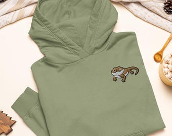 Puede incluir: Una sudadera con capucha de color verde salvia que presenta un gráfico bordado de un dragón barbudo marrón y blanco en el pecho. La sudadera incluye un bolsillo tipo canguro frontal, mangas largas con puños acanalados y capucha sin cordón.