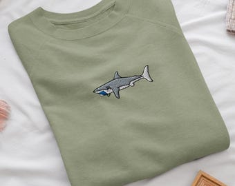 Könnte beinhalten: Ein salbeigrünes Sweatshirt mit Rundhalsausschnitt und einer kleinen, detaillierten Stickerei eines grauen Hais, der horizontal schwimmt. Der Hai ist mit weißem Bauch und einem blauen Akzent am Maul dargestellt und mittig auf der Brust platziert.