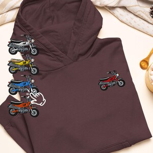 以下が含まれることがあります： 胸に赤いオートバイの刺繍が施されたマルーン色のパーカー。白、黄色、青、オレンジ色の刺繍入りオートバイも表示されています。パーカーにはカンガルーポケットとドローストリングフードが付いています。