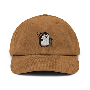 May include: A brown corduroy baseball cap featuring an embroidered graphic of a penguin wearing a top hat and holding a cane. The penguin is black and white with orange accents on the hat and feet, positioned centrally on the front of the cap.