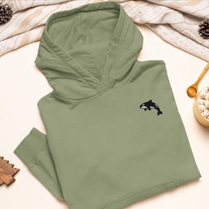 May include: A sage green hooded sweatshirt featuring a small, embroidered black and white orca whale graphic on the upper left chest. The pullover garment includes a hood with a crossover neckline, long sleeves with ribbed cuffs, and a front pocket.