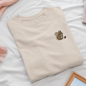 May include: A beige crewneck sweatshirt featuring a small embroidered graphic on the upper left chest. The embroidery depicts a brown squirrel with a bushy tail holding a small brown acorn, rendered in a simple, clean, and detailed style.