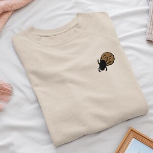 Op de afbeelding: Een beige sweater met ronde hals en een kleine geborduurde afbeelding op de linkerborst. Het borduursel toont een zwarte kever naast een ronde, getextureerde beige vorm die lijkt op een koekje of een maan met een gezicht.