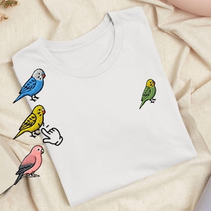 May include: A white short-sleeved t-shirt featuring embroidered designs of four budgerigar birds in blue, yellow, green, and pink, along with a small white hand icon pointing towards the yellow bird on the left side of the garment.
