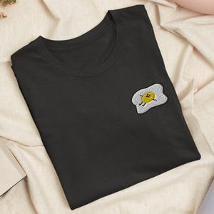 Könnte beinhalten: Ein schwarzes T-Shirt mit kurzen Ärmeln und einem gestickten Aufnäher auf der Brust. Der Aufnäher zeigt ein Spiegelei mit gelbem Eigelb, weißem Eiweißrand und einem kleinen lächelnden Gesicht mit dünnen schwarzen Armen und Beinen.