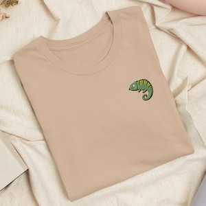 Puede incluir: Una camiseta de cuello redondo en color beige con un pequeño gráfico bordado de un camaleón verde en el lado izquierdo del pecho. El camaleón aparece de perfil con detalles amarillos en el lomo y una cola enroscada, con un diseño sencillo.