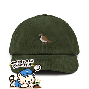 May include: A dark olive green corduroy baseball cap featuring an embroidered brown and white sparrow on the front. In the lower left corner, a cartoon tiger wearing a blue cap looks at a small sprout with text reading WAITING FOR MY DONUT TREE.