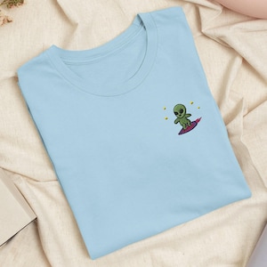 Op de afbeelding: Een lichtblauw t-shirt met ronde hals en een kleine geborduurde afbeelding op de borst. Het borduursel toont een groen alien-figuur dat op een paarse surfplank staat, omringd door verschillende kleine gele sterren op de stof.