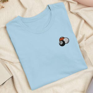 May include: A light blue crew neck t-shirt featuring a small embroidered graphic on the left chest. The embroidery depicts three billiard balls, including a black eight ball, a white ball, and a reddish-brown ball, clustered together in a group.