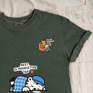 Könnte beinhalten: Ein dunkelolivgrünes T-Shirt mit einer Grafik eines Faultiers, das Kopfhörer trägt und einen Laptop benutzt, mit dem Text JOY OF MISSING OUT, sowie einer Grafik eines Tigers, der unter einer blau karierten Decke schläft, mit dem Text REST IS PRODUCTIVE TOO.