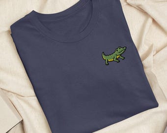 Peut inclure: Un t-shirt à col rond bleu marine foncé avec une petite broderie représentant un alligator vert sur la poitrine gauche. L'alligator est représenté de profil avec un ventre jaune et un détail d'œil noir.