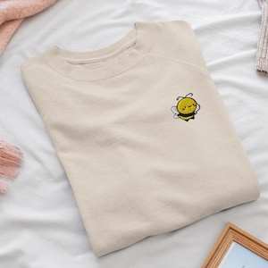 May include: A beige crewneck sweatshirt featuring a small embroidered graphic of a smiling yellow and black bee with white wings on the upper left chest. The garment has long raglan sleeves and a ribbed collar, cuffs, and hemline.