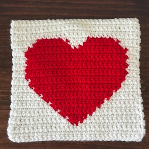 May include: A square, handmade crochet piece featuring a red heart design centered on a white background. The heart is filled with red yarn, and the border is white. The piece is likely a coaster or decorative item.