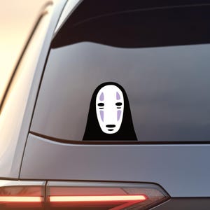 May include: A black vinyl car sticker depicting the character No-Face from the animated film Spirited Away. The sticker shows the character's white mask-like face with purple markings and a black hooded cloak. The sticker is affixed to a car window.