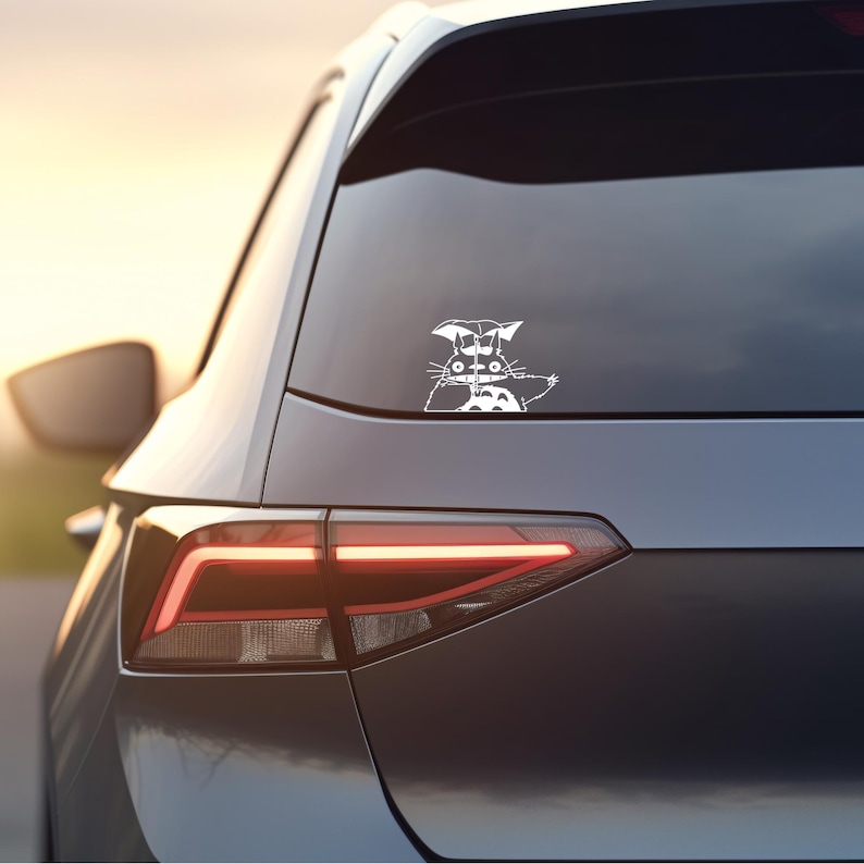 May include: A white vinyl sticker of a cartoon cat with a large head and a small body. The cat is holding an umbrella and is looking to the right. The sticker is on the rear window of a silver car.