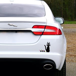 May include: A black cat sticker with large eyes and a curved tail is affixed to the rear of a white car. The sticker includes three small, colorful flower-like shapes. The car has a Jaguar emblem.