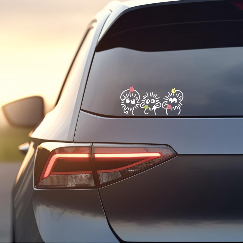 May include: A gray car with a rear window sticker featuring three white cartoon characters with black eyes and colorful flowers on their heads.