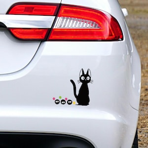 May include: Black cat sticker with a whimsical design, featuring a sitting cat with large eyes and a curved tail, accompanied by three small, round creatures and colorful stars. The sticker is applied to a white car.