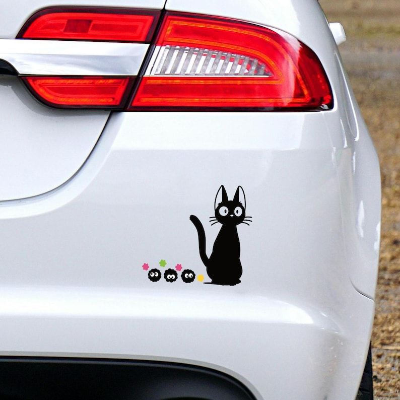 May include: Black cat sticker with a whimsical design, featuring a sitting cat with large eyes and a curved tail, accompanied by three small, round creatures and colorful stars. The sticker is applied to a white car.