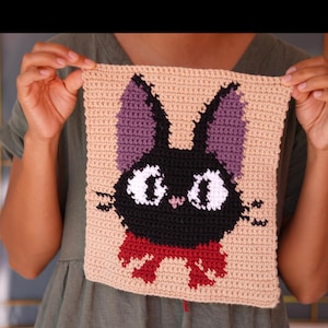 May include: A square crochet piece featuring a black cat design with large white eyes, purple inner ears, and a red bow tie. The cat is centered on a light beige background, created with visible yarn stitches in a grid-like pattern.
