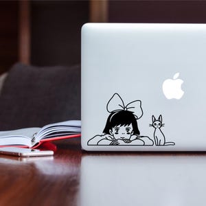 Könnte beinhalten: Ein schwarz-weißer Aufkleber mit einem Mädchen mit Schleife im Haar und einer Katze, die daneben sitzt. Der Aufkleber befindet sich auf einem silbernen Laptop.