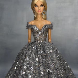 May include: A fashion doll with long blonde hair wearing a silver tiara with pink stones, dangling earrings, and an off-the-shoulder ball gown covered in silver sequins and metallic circular embellishments over a dark grey mesh fabric.