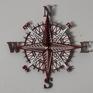 May include: A wall clock shaped like a compass rose featuring layered wood in dark brown, white, and deep red. The clock face displays cardinal directions N, S, E, and W, along with intermediate points, with thin clock hands centered on the design.