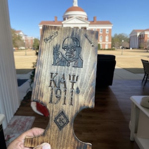 May include: A wooden plaque with a dark brown finish, featuring an engraved portrait of a man, the Greek letters "ΚΑΨ", and the year "1911". The plaque also includes the text "Ella Mae Day" and "Smooth G".