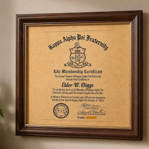 Könnte beinhalten: Gerahmte Kappa Alpha Psi Fraternity Life Membership Certificate. Das Zertifikat mit Text befindet sich in einem dunkelbraunen Holzrahmen. Das Zertifikat ist beige mit schwarzem Text und einem Bruderschaftswappen.