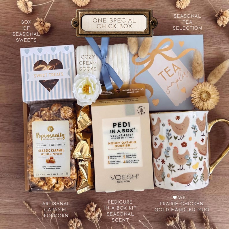 Op de afbeelding: Een geschenkdoos met een zak Popinsanity karamel popcorn, een Voesh Pedi in a Box set, een witte mok met kippenpatroon en gouden oor, een doosje Sweet Treats, een doosje Tea Sampler en cr&egrave;mekleurige sokken vastgebonden met een blauw lint.