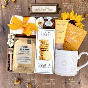 May include: A gift box labeled A Little Sunshine Box containing cream socks with a yellow ribbon, a jar of honey, a tube of Deep Steep grapefruit bergamot lotion, a box of Cup of Sunshine herbal tea, a box of Rustic Bakery vanilla shortbread, a Pedi in a Box kit, a wooden honey dipper, and a white mug with a daisy pattern.
