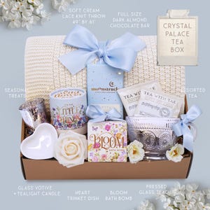 May include: A gift basket with a soft cream lace knit throw (49" x 61"), a dark almond chocolate bar, and a Crystal Palace tea box. It includes a glass votive with a tealight candle, a heart trinket dish, a Bloom bath bomb, and assorted tea.