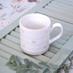 May include: A white ceramic mug featuring a ribbed texture and small, light-colored heart patterns scattered across the exterior. The mug has a curved handle and a smooth, unglazed base, showcasing a simple design with a clean, rounded silhouette.